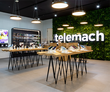 Različni pametni telefoni in dodatki so razporejeni na svetlo rjavih lesenih mizah s črnimi kovinskimi nosilci. Velik, stiliziran napis Telemach je viden ob steni iz umetnega zelenja. Trgovina ima svetel, sodoben dizajn.