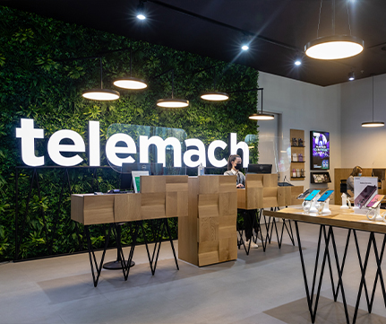 Notranjost trgovine Telemach. Sodoben, svetel prodajni prostor z velikim, osvetljenim napisom Telemach na steni z zelenjem. Vidno je razstavljen svetlo rjav lesen recepcijski pult, na njem pa so razstavljeni različni mobilni telefoni.