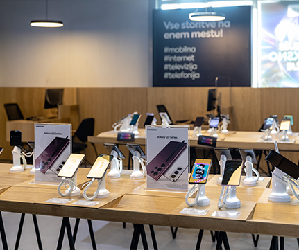 Sodoben razstavni prostor pametnih telefonov v dobro osvetljeni trgovini prikazuje mobilne telefone, razstavljene na lesenih mizah z varnostnimi stojali, s poudarkom na najnovejši tehnologiji in funkcijah fotoaparata.